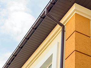 Gutter-Installation-Kentwood-MI-Michigan-2 | JM Roofing Solutions Close-up of a yellow-orange building corner with a white-framed window and a brown gutter along the roof edge.