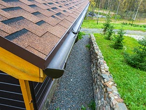 Gutter-Repairs-Kentwood-MI-Michigan-1 | JM Roofing Solutions Close-up of a brown shingled roof edge with a black gutter and downspout, next to a gravel path, stone wall, and grassy yard.