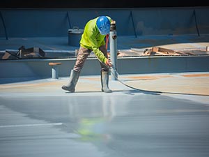 Roof-Coating-Kentwood-MI-Michigan-1 | JM Roofing Solutions Construction worker in a neon safety vest and blue helmet smoothing an epoxy coating on a wide concrete floor with a long-handled tool.